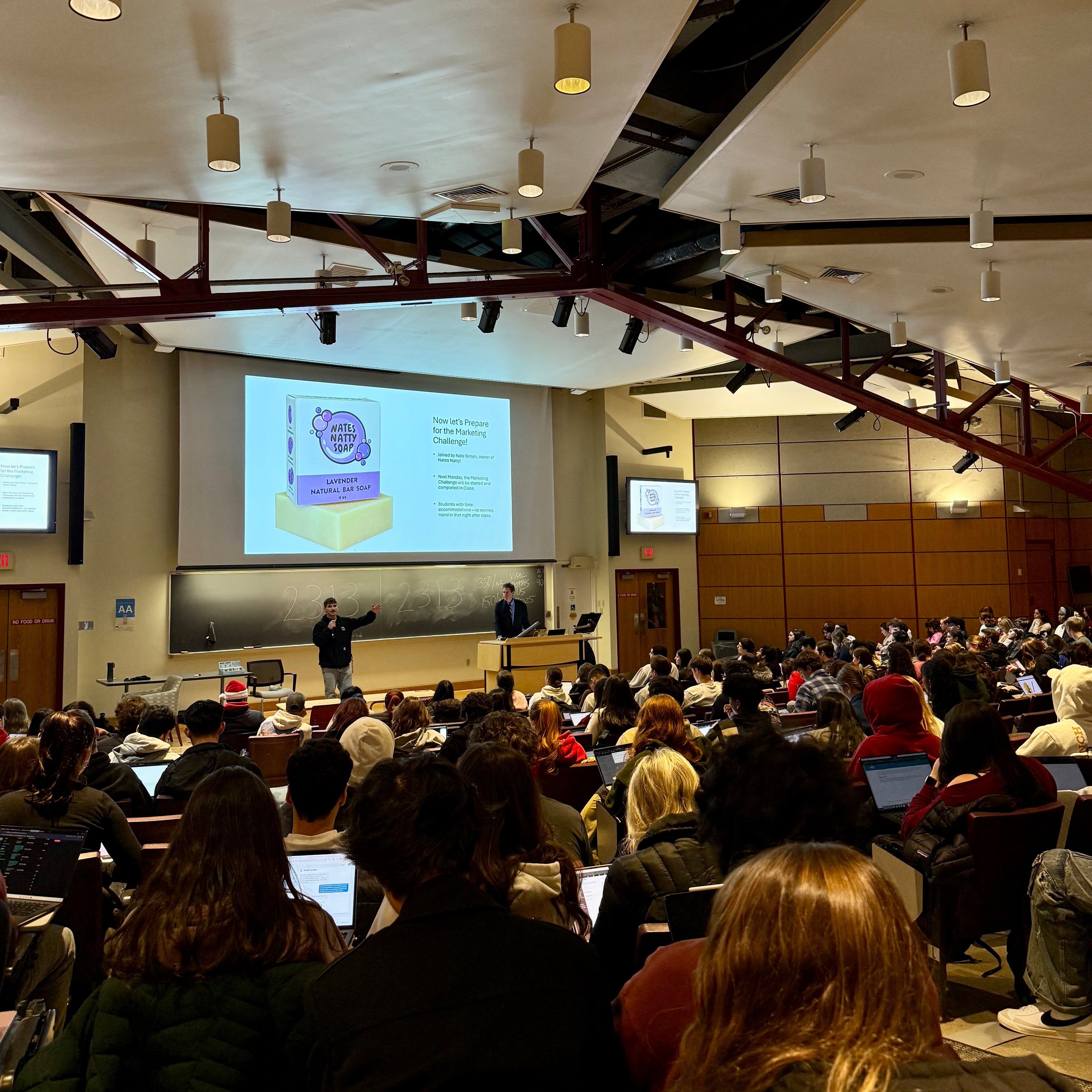Lecture hall with a lecture in progress, audience facing the front where a presenter is standing.